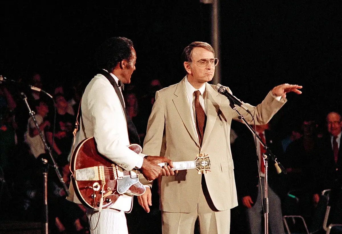 Photographie : sur une scène, Chuck Berry, guitare en main et costume blanc, à côté de Carl Sagan qui tend le bras vers le public.
