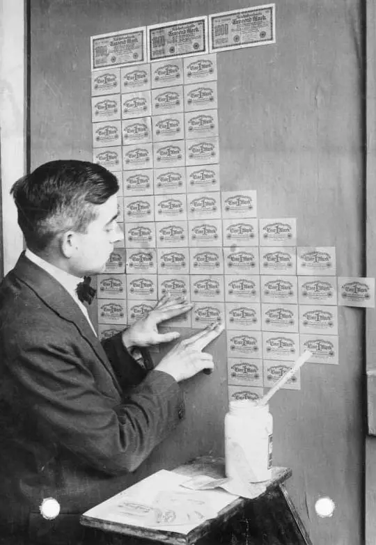 Photographie en noir et blanc : un homme colle des billets de banque sur un mur, comme du papier peint.