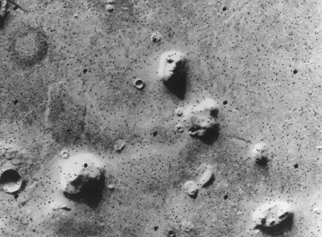 Photographie en noir et blanc de la surface de Mars : au milieu de collines, l'une ressemble à un visage humain vu de face, avec des yeux, un nez et une bouche.