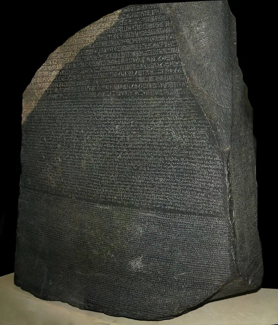 Photographie : une stèle de pierre sombre, brisée en haut, couverte de trois textes gravés l'un sous l'autre.