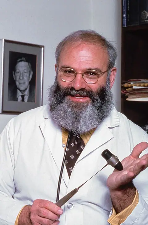 Photographie : Oliver Sacks, barbe grise et blouse blanche, sourit en tenant un marteau à réflexes et un diapason.