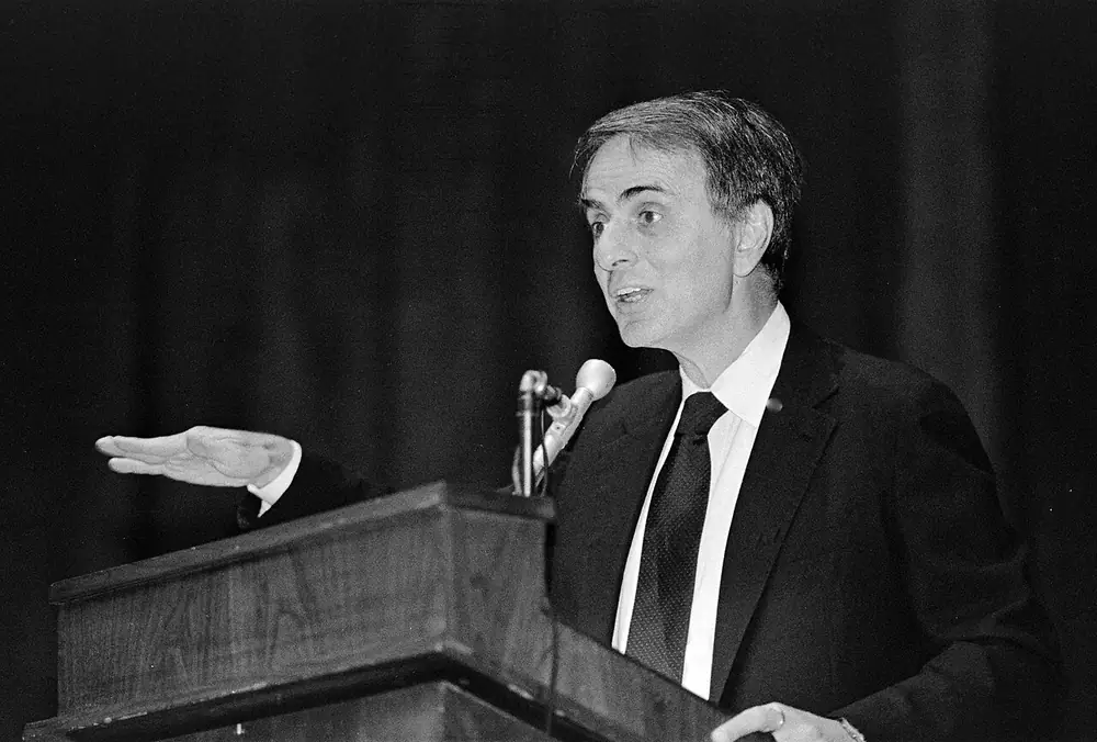 Photographie en noir et blanc : Carl Sagan parle derrière un pupitre, la main tendue vers le public.