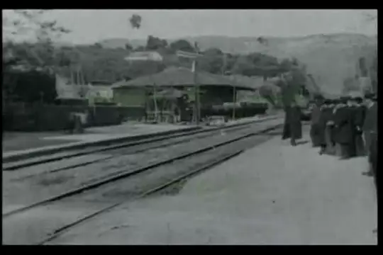 Film muet en noir et blanc : une locomotive à vapeur arrive de loin vers la caméra, le long d'un quai où attendent des voyageurs, puis s'arrête.